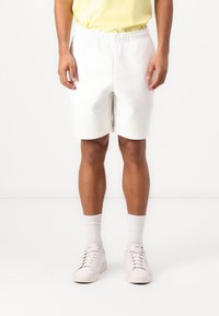Hvite shorts til kneet med elastisk midje, kombinert med hvite sokker og sneakers, mot en ensfarget bakgrunn.