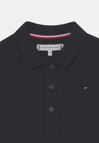 Námořnická modrá polo košile s vroubkovaným límečkem, dvouknoflíkovou légou a výrazným výšivkovým logem v červené, bílé a modré na hrudi.