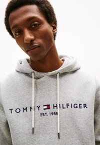 Joven con cabello corto y rizado que lleva una sudadera con capucha gris claro con "Tommy Hilfiger Est. 1985" bordado en el frente.
