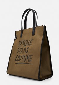 Brązowa torba na ramię z płótna z czarnymi wykończeniami i uchwytami, z wyraźnym napisem "VERSACE JEANS COUTURE" z przodu. Zamek błyskawiczny na górze.