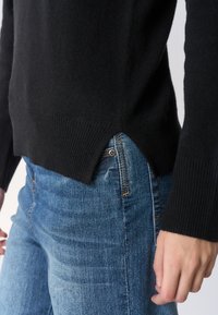 Maglione nero in maglia con polsini e orlo a coste, con spacco laterale. Abbinato a jeans di denim blu. La texture appare morbida e accogliente.