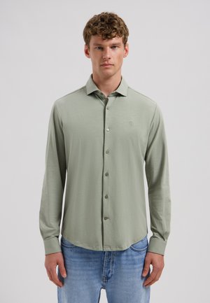 Junger Mann mit lockigem Haar, der ein hellgrünes, langärmliges Hemd mit Knöpfen und blaue Jeans trägt und vor einem schlichten hellen Hintergrund steht.