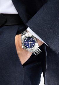 Orologio da polso in metallo argentato con quadrante blu, funzione data e bracciale tono argento, indossato sopra un giubbotto di giacca blu navy con una camicia bianca.
