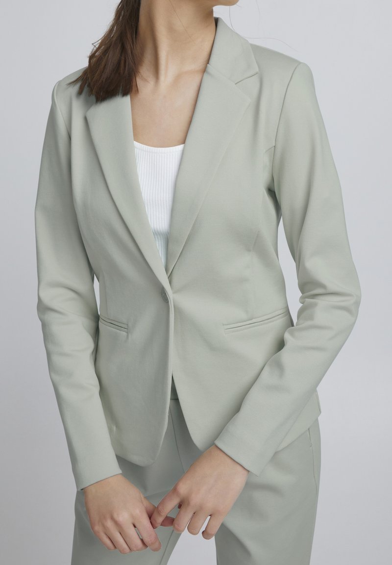 Femme portant un blazer gris clair ajusté et un pantalon assorti, avec un top blanc côtelé en dessous, debout devant un fond gris.