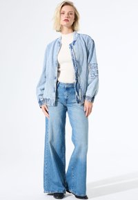 Femme blonde debout portant une veste en denim bleu clair, un jean évasé, un haut à côtes blanc et des chaussures noires sur fond blanc.