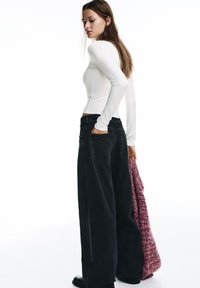 Witte top met lange mouwen gedragen met zwarte, wijd uitlopende jeans met hoge taille. Model houdt een roze gebreide trui met textuur vast, staande tegen een witte achtergrond.