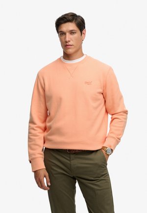 Hombre con sudadera color melocotón con cuello redondo, camiseta interior blanca, pantalones verde oliva, cinturón marrón y reloj de pulsera, de pie frente a fondo blanco.