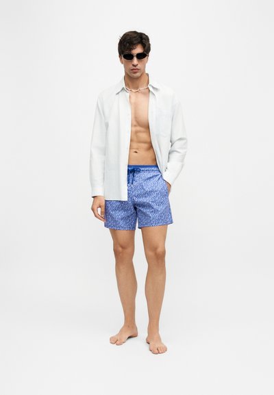 Jeune homme portant une chemise blanche ouverte, un short de bain bleu à motifs, des lunettes de soleil et un collier de coquillages, debout pieds nus devant un fond uni.