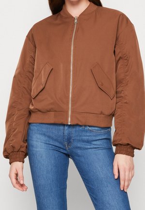 Mujer con chaqueta bomber marrón con cremallera, bolsillos con solapas y puños fruncidos, combinada con vaqueros azules de denim.