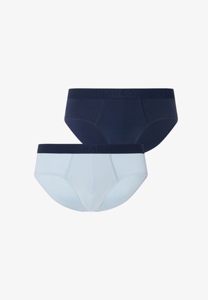 Mand med kort mørkt hår og skæg iført marineblå underbukser, stående barfodet mod en ensfarvet lys grå baggrund, kiggende sidelæns.