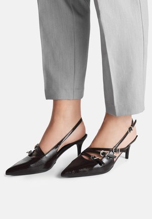 Zapatos de salón con tacón bajo tipo kitten, de charol negro con tiras traseras, puntera puntiaguda y múltiples hebillas plateadas, usados con pantalones cortos de color gris claro.