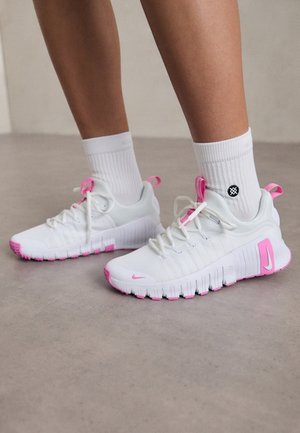 Ben iført hvide sneakers med pink accenter og hvide mellemlange sokker, stående på gråt gulv.