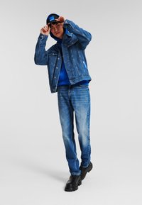 Jachetă din denim cu buzunare pe piept; hanorac albastru dedesubt; blugi albaștri decolorati; cizme negre; persoană care își ajustează o șapcă. Logo alb pe jachetă.