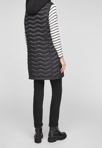 Schwarze gepolsterte Weste mit einem gewellten Texturmuster, über einem schwarz-weißen gestreiften Langarmshirt getragen, kombiniert mit schwarzen Jeans und Stiefeln.