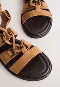 Sandales en cuir marron avec des brides en daim beige et des œillets métalliques, avec un design à lacets et une semelle intérieure rembourrée.