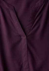 Tessuto color prugna con una consistenza liscia, caratterizzato da un design a V e cuciture precise lungo i bordi, con particolare attenzione alla zona del collo.