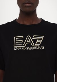 Black t-shirt featuring a large, contrasting cream-colored graphic that reads "EA7 EMPORIO ARMANI." Short sleeves and round neckline.