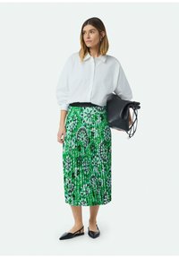 Witte blouse met pofmouwen, groene plissérok met bloemenpatronen, en zwarte slingback flats. Zwarte handtas in de hand.