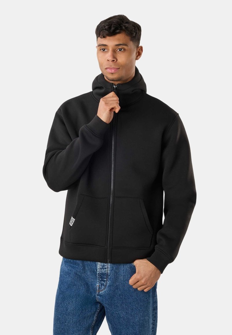 Jeune homme portant un sweat à capuche noir zippé avec poches kangourou et jean bleu, tirant la fermeture éclair près de son cou.