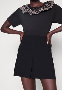 Personne portant une robe noire à manches courtes avec un col festonné brodé de fleurs et des détails de boutons sur les côtés de la jupe.