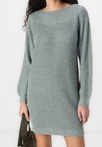 Femme portant une robe-pull en maille grise texturée à manches longues, tenant un sac en velours côtelé vert foncé sur un fond blanc uni.