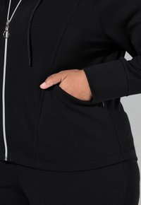 Černá sportovní bunda má celopropínací zip, kapuci se stahovací šňůrkou a boční kapsy. Je vyrobena z hladkého, měkkého materiálu a má přiléhavý střih.