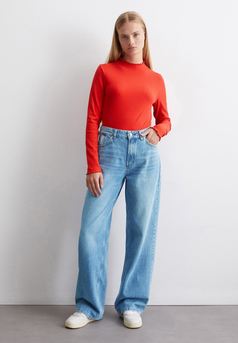 Rotes gestreiftes Langarmtop mit einem gewellten Ausschnitt, kombiniert mit hellblauen weit geschnittenen Jeans und weißen Sneakers, vor einem einfarbigen Hintergrund.