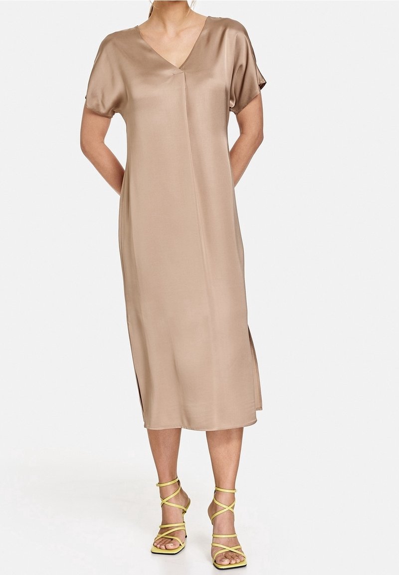 Femme portant une robe midi en satin beige à manches courtes et des sandales à talons hauts jaunes à brides, debout, les mains dans le dos.