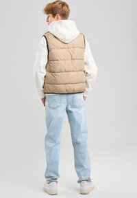 Beige peitto liivi pysty saumoilla, huppari valkoisena alla, vaaleansiniset farkut ja valkoiset lenkkarit, nähtynä takaa.