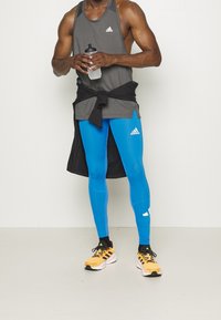 Mužský atleta v šedém tílku Adidas, jasně modrých legínách Adidas, žlutých běžeckých botách Adidas, držící lahev s vodou, s černou bundou uvázanou kolem pasu.