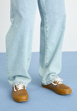 Baskets en daim marron avec des accents blancs, des semelles en caoutchouc et des lacets blancs ; associées à un jean en denim bleu clair, légèrement ample.