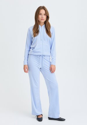 Jeune femme aux longs cheveux portant un sweat à capuche zippé bleu clair et un pantalon assorti, debout droite sur un fond clair uni.