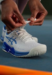 Balti sportiniai batai su mėlynais akcentais, turi tinklinį viršų, tekstūruotą pado paviršių ir matomą logotipą. Kengūros yra rišamos.