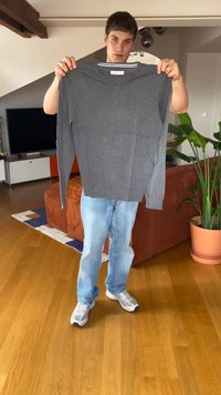 Junge Person in Jeans und Sneakers, die einen grauen V-Ausschnitt-Pullover in einem Wohnzimmer mit Holzboden und Pflanzen hält.