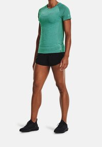 Camiseta atlética verde de mangas cortas con detalles texturizados, combinada con pantalones cortos negros y zapatillas negras. La camiseta presenta un diseño ajustado.
