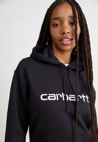 Felpa con cappuccio nera con logo "carhartt" ricamato in bianco, caratterizzata da una texture morbida e una vestibilità rilassata. Capelli intrecciati visibili.