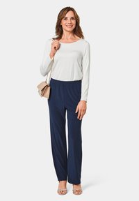 Witte blouse met lange mouwen, marineblauwe broek met elastische tailleband en beige sandalen met bandjes. Kleine beige handtas met kettinghengsel. Gladde stoftexturen.