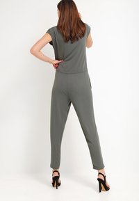 Oliwkowy kombinezon z krótkimi rękawami, zwężanymi nogawkami i szwem w talii. Noszony z czarnymi sandałami na wysokim obcasie. Gładka tekstura materiału.