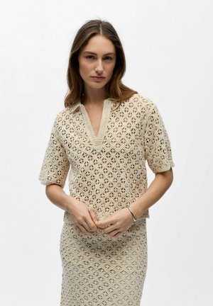 Kvinne iført beige heklet topp med V-hals og matchende skjørt, stående med hendene foldet foran hvit bakgrunn.