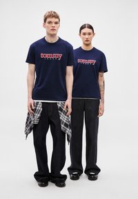 Camisetas de algodón navy con el logo "tommy" en rojo y el texto "JEANS". El modelo lleva vaqueros negros de pierna ancha con una camisa de cuadros anudada a la cintura.
