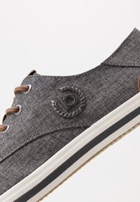 Baskets en tissu gris avec une surface texturée, des lacets marron et un logo métallique sur le côté. Comprend une semelle intermédiaire blanche et une semelle extérieure en caoutchouc.