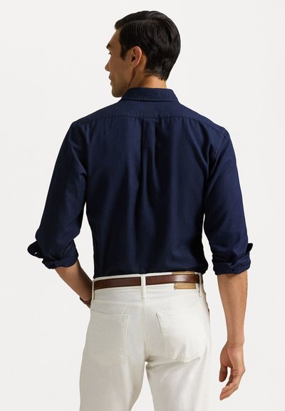 Cămașă albastru închis cu mâneci lungi și manșete răsucite, cu o linie dreaptă la spate, asociată cu pantaloni albi și o curea de piele maro. Design simplu, fără modele.