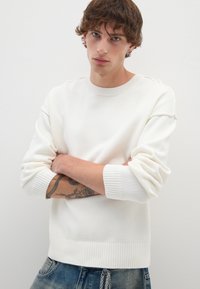 Witte gebreide trui met ribgebreide manchetten en zoom, voorzien van een ronde halslijn en subtiele stikdetails. Model draagt een blauwe spijkerbroek.