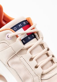Sneaker beige con una toma in pelle e rete testurizzata, accenti arancioni e una linguetta blu navy con una striscia di logo rossa e bianca.