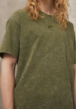 Persoon met krullend blond haar die een olijfgroen Nike T-shirt draagt met een subtiele gemarmerde structuur en een klein geborduurd logo.