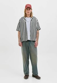 Chemise à manches courtes à rayures noires et blanches, t-shirt gris, jeans bleu délavé, chaussures marron, casquette rouge avec inscription, tatouages sur les bras.