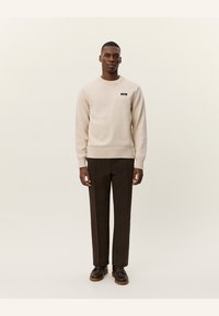 Jersey beige de punto con cuello redondo, dobladillo acanalado y un pequeño parche negro con logo. Combinado con pantalones rectos marrones y zapatos marrones.