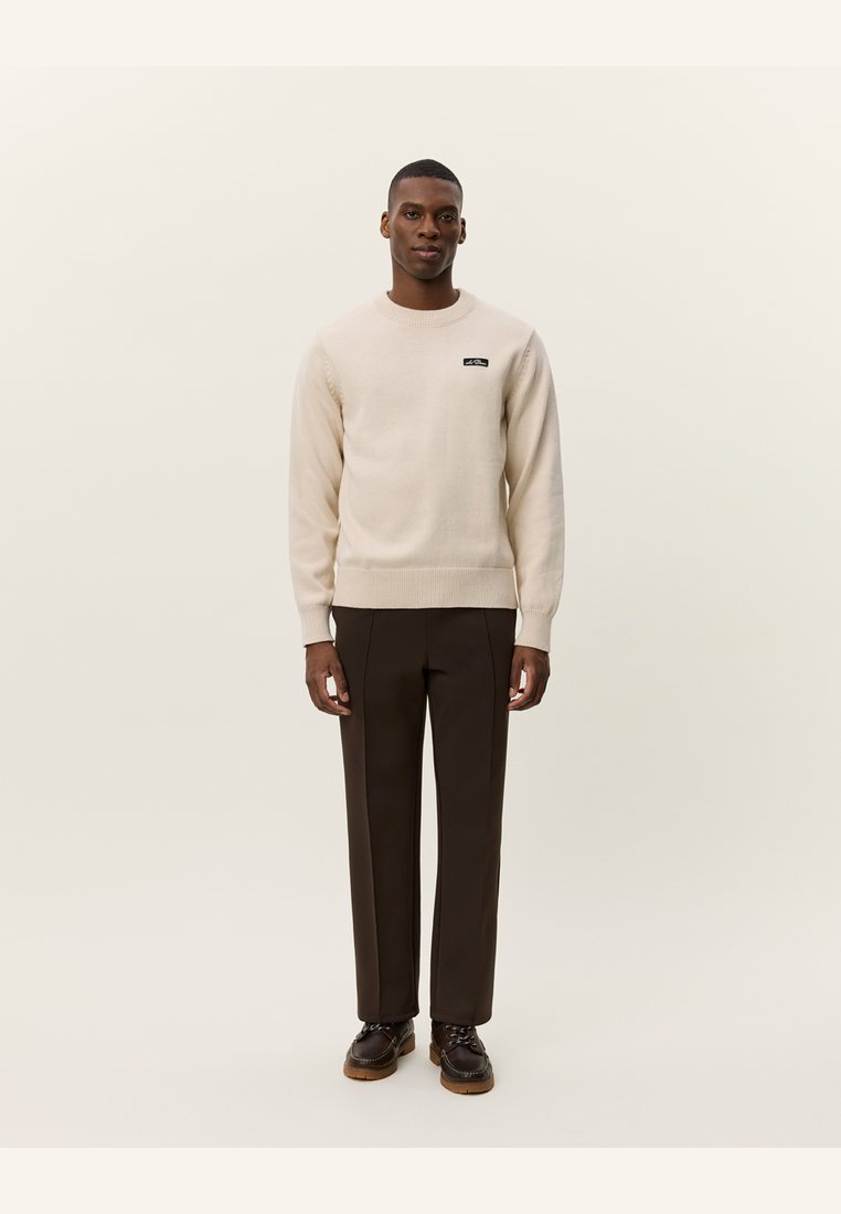 Jersey beige de punto con cuello redondo, dobladillo acanalado y un pequeño parche negro con logo. Combinado con pantalones rectos marrones y zapatos marrones.