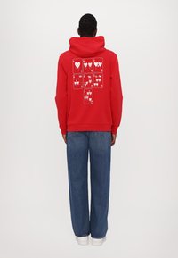 Red hoodie with a large graphic of white hearts and cards on the back. Features a kangaroo pocket and drawstring hood. Paired with blue jeans.