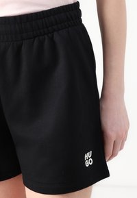 Black shorts made of soft fabric with an elastic waistband. Features small white "HUGO" logo on the lower left. Simple design, no patterns.